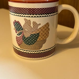 May include: A white ceramic mug with a red rim and a patchwork-style design of a brown and green chicken. The mug has a red, white and green border with a white polka dot pattern.