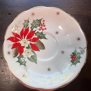 May include: White saucer with a gold rim and a festive Christmas design. The design features a red poinsettia, green holly leaves, red berries, and small gold stars. The saucer has a scalloped edge.