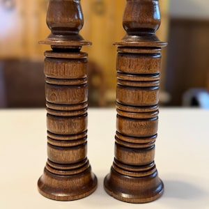 May include: Two wooden candle holders with a dark brown finish. Each holder has a rounded base, a stacked cylindrical body, and a flared top. The candle holders are displayed on a white surface.