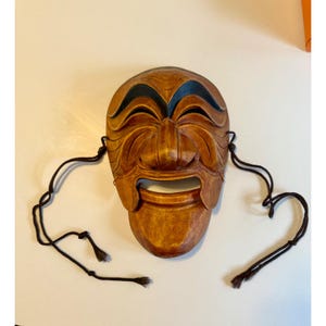 Puede incluir: Una máscara de madera con una cara sonriente, de color marrón claro con detalles marrón oscuro alrededor de los ojos y la boca. La máscara tiene un diseño tallado y cuerdas negras. Objeto cultural.