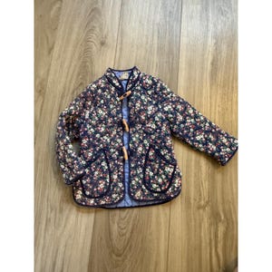 May include: A navy blue quilted jacket with a floral pattern in pink, white, and green. The jacket has a round collar, wooden toggle closures, and two front pockets. The trim is a darker navy blue.