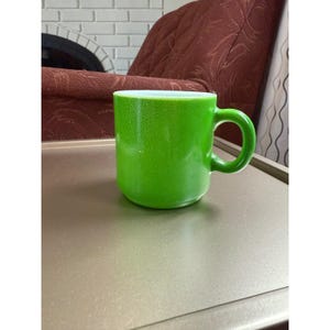 Könnte beinhalten: Eine leuchtend grüne Tasse mit rundem Korpus und gebogenem Henkel. Die Tasse hat eine strukturierte Oberfläche und einen weißen Innenraum. Sie steht auf einer hellbraunen Oberfläche.