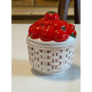 May include: A ceramic trinket box with a basket weave design. The lid is topped with a cluster of red, glossy strawberries with green accents. The box is white with a light brown trim.