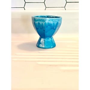 May include: A small, turquoise ceramic cup with a pedestal base. The cup has a hexagonal shape and a glossy finish with white drips around the rim. The cup is displayed on a light pink surface.