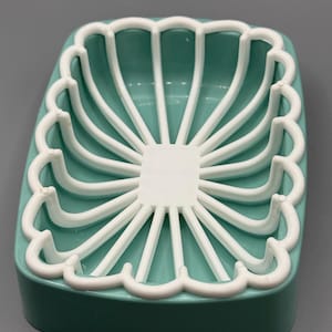 May include: A teal soap dish with a white, scalloped, fan-like design. The dish has a rectangular shape with rounded edges and a raised rim. The white design is centered on the teal base.