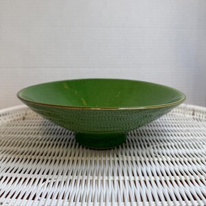 May include: A green ceramic bowl with a textured exterior and a gold rim. The bowl has a wide, shallow design and sits on a white wicker surface. The bowl is a vibrant shade of green.