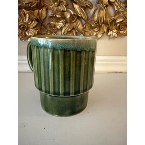 May include: A green ceramic mug with vertical ribbed detailing. The mug has a handle and a two-tiered design, with a darker green base and a lighter green rim. The mug is likely vintage or retro.