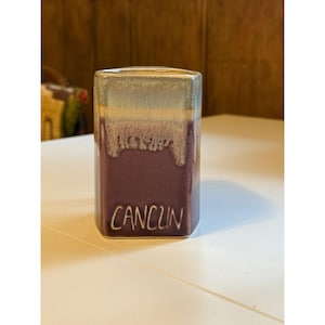 Puede incluir: Un jarrón rectangular de cerámica con un acabado degradado. La parte superior es de color gris-beige claro, que pasa a una banda blanca texturizada y luego a una base de color púrpura intenso. La palabra "CANCUN" está escrita en blanco en la parte delantera.