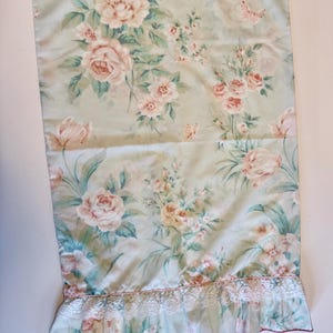 Könnte beinhalten: Floraler Stoff mit hellgrünem Hintergrund und einem Muster aus rosa Rosen und grünen Blättern. Der untere Rand hat eine weiße Spitzenborte mit rotem Rand. Geeignet für Heimdekoration.