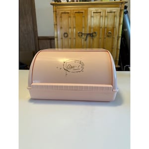 May include: A vintage pink plastic bread box with a curved top and a rectangular base. The box has a red trim and a decorative gold-colored graphic that says "Bee-". The bread box is on a white surface.