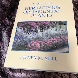Puede incluir: Un libro titulado "Manual of Herbaceous Ornamental Plants" de Steven M. Still. La portada presenta una fotografía de un macizo de jardín con plantas moradas, rosas y plateadas. El libro tiene una cubierta color crema con texto azul.