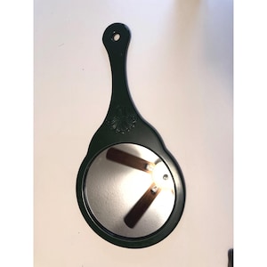 May include: A dark green hand mirror with a round mirror and a long handle. The handle has a decorative shell design. The mirror reflects a ceiling fan.