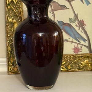 May include: A dark burgundy glass vase with a clear glass base. The vase has a rounded body and a narrow neck with a flared rim. The background includes a framed botanical print with birds.