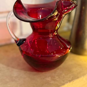 Puede incluir: Una jarra de vidrio rojo rubí con un asa transparente y un borde acampanado. La jarra tiene una base redondeada y una superficie lisa y brillante. Probablemente se utiliza para servir bebidas.