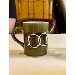 May include: Olive green ceramic mug with a curved handle. The mug features a white and brown chain-link design around the middle. The mug is sitting on a white surface.