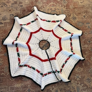May include: A handmade, white crochet Christmas tree skirt with red and green accents. The skirt has a black border and a circular opening in the center. The design features a starburst pattern.