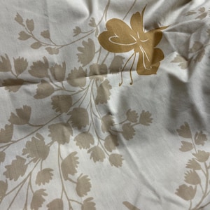 Puede incluir: Tela color crema con un estampado floral repetido en tonos beige y dorado. El diseño presenta flores estilizadas y ramas con hojas, creando una estética delicada y natural. La tela parece suave y ligeramente arrugada.