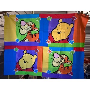 Könnte beinhalten: Ein farbenfrohes Stoffpaneel mit Winnie Puuh und Tigger. Das Panel zeigt vier Quadrate mit Bildern von Winnie Puuh und Tigger, umgeben von Blumen und Schmetterlingen. Die Hintergrundfarben sind Gelb, Grün, Blau, Lila und Orange.