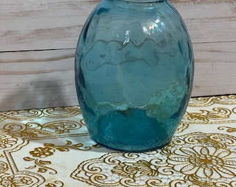 Puede incluir: Un jarrón de vidrio azul claro con una superficie texturizada. El jarrón tiene forma ovalada y una abertura ancha. El jarrón está sobre una superficie con estampado blanco y dorado. El jarrón mide unos 20 cm de alto.