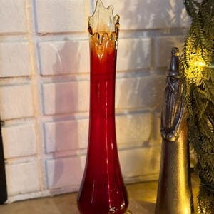 May include: A tall, slender red glass vase with a flared, scalloped rim. The vase has a clear, amber-coloured top and rests on three small, clear feet. A decorative item is next to the vase.