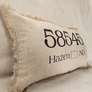 May include: A beige pillow with a frayed edge features the text "58545 Hazen ND" printed in black.