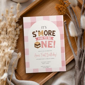 Może przedstawiać: Prostokątne zaproszenie na urodziny z różowo-białym tłem w kratkę vichy i łukowatym środkiem. Tekst brzmi IT'S S'MORE FUN TO BE ONE! z grafiką s'more, a poniżej znajdują się szczegóły pierwszych urodzin Avy w dniu 11 lipca 2026 roku.