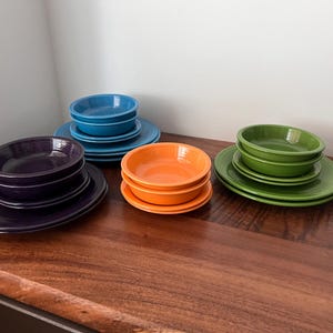 May include: A set of four stackable ceramic bowls in different colours, including purple, orange, green, and blue. The bowls are arranged in three stacks on a wooden surface.