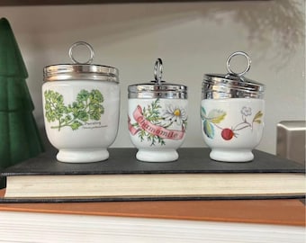 Pode incluir: Três ovos de cerâmica branca com tampas prateadas e desenhos florais. Os ovos são etiquetados como "Salsa", "Camomila" e "Morango".