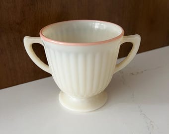 Könnte beinhalten: Eine weiße Milchglasschüssel mit rosa Rand und geriffelten Seiten. Die Schüssel hat zwei Henkel und einen Sockel.