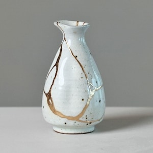 May include: A small, pear-shaped ceramic vase with a white, speckled glaze. The surface features prominent, irregular gold-colored lines that mimic the appearance of repaired cracks, extending from the narrow neck down to the wider, rounded base.