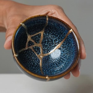 May include: A small, round ceramic bowl featuring a deep blue, textured glaze with a dark brown rim. The bowl displays a prominent kintsugi repair, where the cracks are filled with a distinct, reflective gold-coloured lacquer finish.