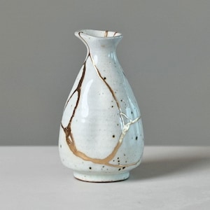 May include: A small, pear-shaped ceramic vase with a white, speckled glaze. The surface features prominent, irregular gold-colored lines that trace across the body, creating a distinct pattern of interconnected segments around the vessel.