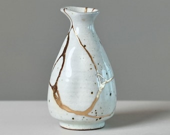 May include: A small, pear-shaped ceramic vase with a white, speckled glaze. The surface features prominent, irregular gold-coloured lines that trace across the body, creating a distinct pattern of interconnected segments around the vessel.