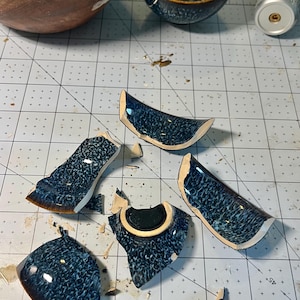May include: Five broken ceramic shards from a bowl with a dark blue and black mottled glaze finish. The pieces show a smooth, glossy exterior surface and a light-coloured clay interior. The shards are scattered on a white cutting mat grid.