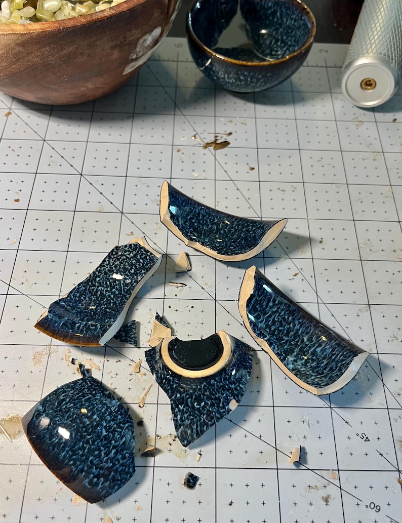 May include: Five broken ceramic shards from a bowl with a dark blue and black mottled glaze finish. The pieces show a smooth, glossy exterior surface and a light-coloured clay interior. The shards are scattered on a white cutting mat grid.