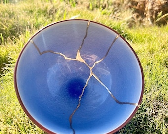 May include: A circular bowl with a smooth, light blue interior that transitions to a deeper blue at the centre. The bowl features a dark brown rim and is repaired with gold-coloured lacquer lines that create a branching pattern across the surface.
