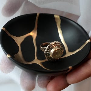May include: A small, round, matte black ceramic dish featuring gold-colored veins that mimic a repaired surface. Resting in the center of the dish is a silver-toned ring with an intricate, textured band and a central circular stone setting.