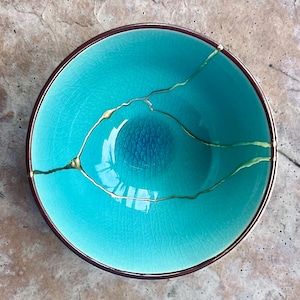 May include: A round ceramic bowl with a vibrant turquoise crackle glaze finish. The bowl features a dark brown rim and gold-colored repair lines running across the interior surface, highlighting the fractured ceramic pieces joined together.