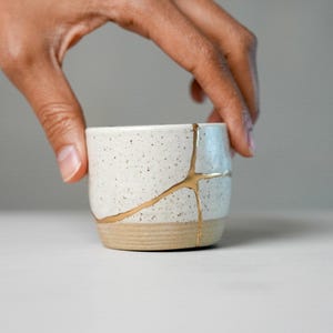 May include: A small, speckled white ceramic cup featuring a gold-coloured repair line running across its surface. The base of the cup is unglazed, showing a natural tan clay texture, while the upper portion has a smooth, speckled white finish.