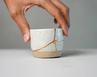 May include: A small, cylindrical ceramic cup with a speckled white glaze on the upper portion and a smooth, unglazed tan base. The cup features a prominent, jagged crack running across the side, which is filled with a metallic gold-coloured material.