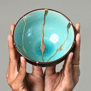 May include: A round ceramic bowl featuring a vibrant turquoise interior with a fine crackle glaze finish. The bowl is decorated with prominent gold-colored lines that extend from the rim toward the center, creating a distinct repair-like pattern.
