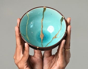 May include: A round ceramic bowl featuring a vibrant turquoise interior with a fine crackle glaze finish. The bowl is decorated with prominent gold-colored lines that extend from the rim toward the center, creating a distinct repair-like pattern.