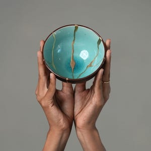 May include: A round ceramic bowl featuring a vibrant turquoise interior with a fine crackle glaze finish. The bowl is decorated with prominent gold-colored lines that traverse the surface, creating a distinct pattern across the basin.