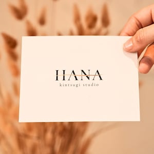 May include: A rectangular white card featuring the black text HANA with a horizontal gold line running through the centre of the letters. Below the name, the words kintsugi studio are printed in a smaller, simple black sans-serif font.