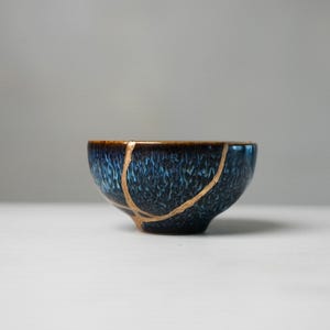 May include: A small ceramic bowl featuring a deep blue and black mottled glaze with a textured finish. The bowl displays a prominent repair line running across its surface, highlighted with a metallic gold-coloured lacquer finish.