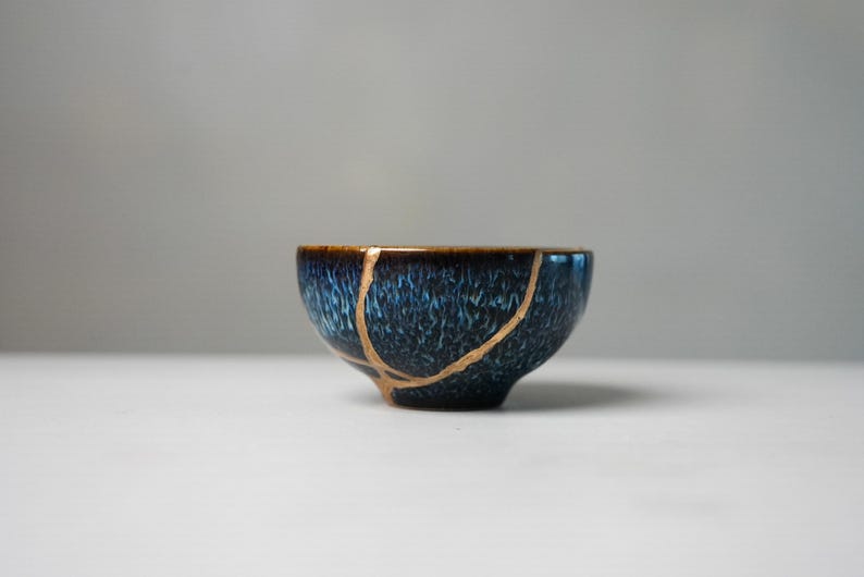 May include: A small ceramic bowl featuring a deep blue and black mottled glaze with a textured finish. The bowl displays a prominent repair line running across its surface, highlighted with a metallic gold-coloured lacquer finish.