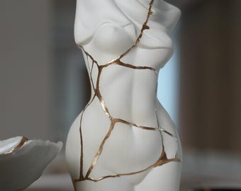 May include: A white ceramic torso sculpture featuring a draped fabric effect across the upper chest. The surface is segmented by thin, metallic gold lines that trace irregular cracks across the entire form, highlighting the contours of the body.