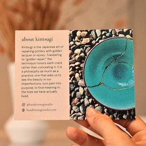 May include: A rectangular informational card with a light peach background on the left featuring black text about kintsugi, and a photograph on the right showing a turquoise bowl repaired with gold lacquer, resting on a bed of small grey stones.