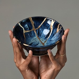 May include: A deep blue ceramic bowl featuring a kintsugi design with gold-colored lacquer filling the cracks across the surface. The bowl has a glossy finish with variations in blue tones, ranging from dark navy to lighter shades of blue.