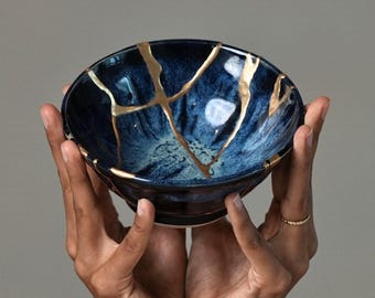May include: A deep blue ceramic bowl featuring a kintsugi design with gold-coloured lacquer filling the cracks across the surface. The bowl has a glossy finish with variations in blue tones, ranging from dark navy to lighter shades of blue.